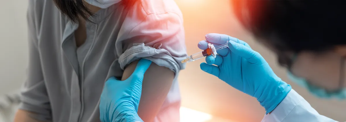 Image of hands in blue medical gloves administering a vaccine into the arm of a patient. The patient's shirt sleeve is rolled up.