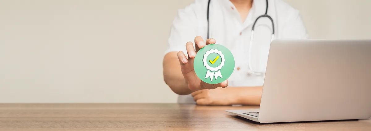 Clinician sitting at table with laptop holding up a checkmark badge