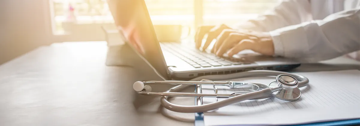 Doctor at desk on laptop with notebook beside it