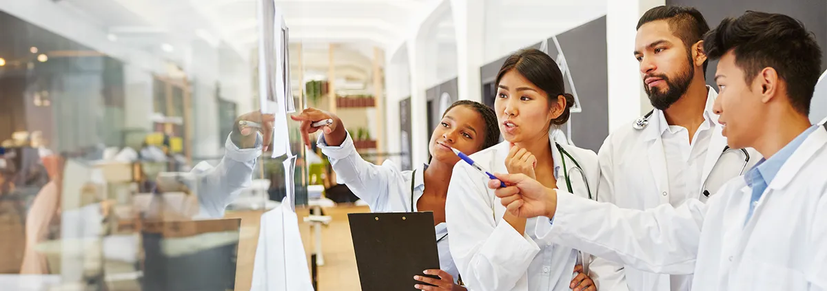 Image of a diverse group of doctors grouped together on the right pointing to and looking at a clear dry erase board on the left.