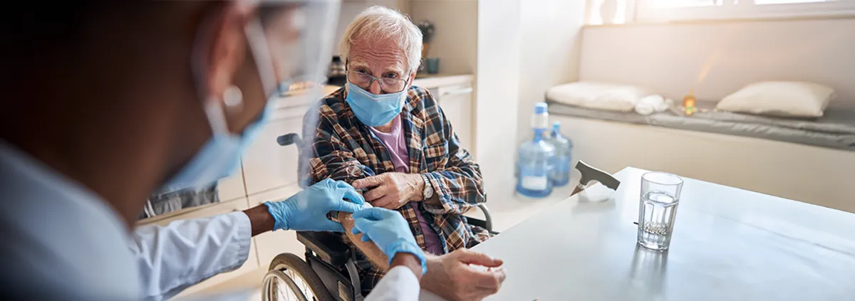 Image of older man wearing a medical mask and sitting in a wheel chair being attended to by a medical professional wearing am medical mask.