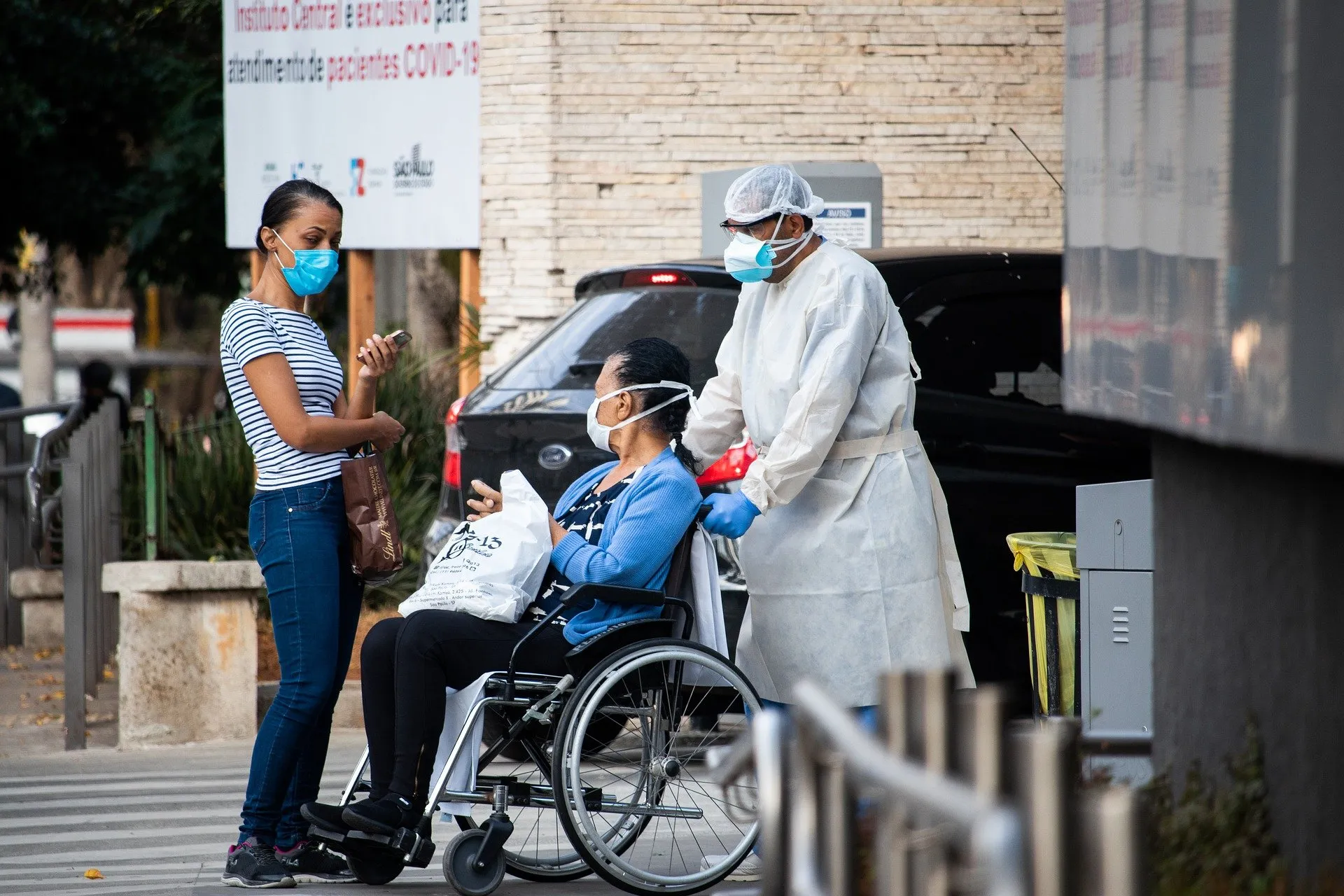 Healthcare professional escorting patient in wheelchair, while patient interacts with woman with mask on.