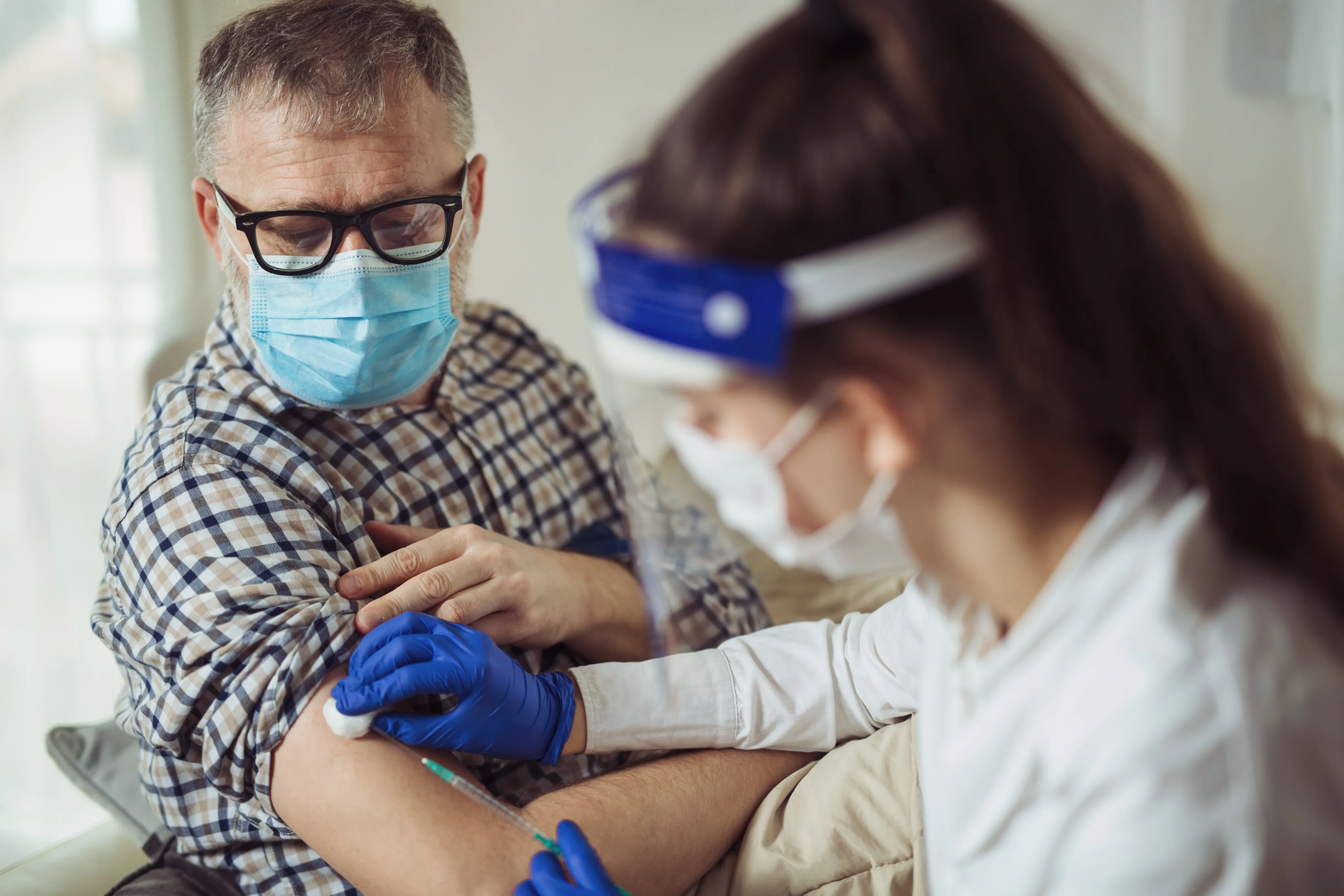 Young woman nurse with surgical mask and face shield giving injection to senior man at home or in a nursing home. 