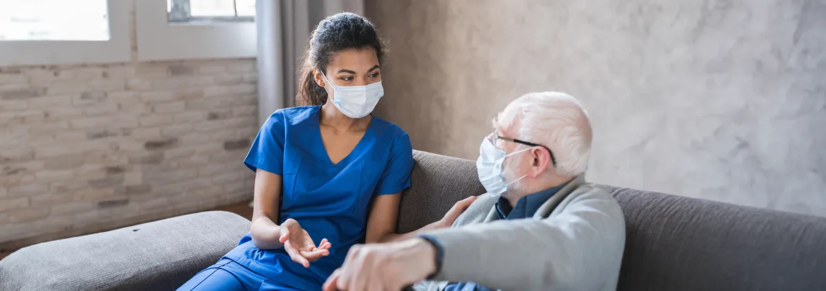 Doctor or female caregiver with senior man grandfather wearing protective mask to protect from Covid 19 at home or nursing home. Doctor talking consulting elderly patient at home